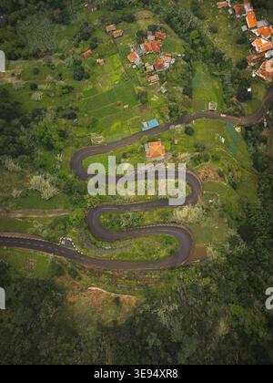 Blick aus der Vogelperspektive auf die gewundene Serpentinenstraße durch üppige grüne Hügel, Terrassenfelder und Häuser mit orangefarbenen Dächern in Ribeiro Frio, Madeira, Portugal. Stockfoto
