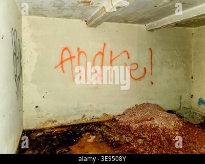 Orange Graffiti bedeutet „Andre!“ An einer schmutzigen, zerrissenen Wand. Der matschige Boden zeigt Anzeichen von Vernachlässigung und Verfall. Deckenbalken zeigen Verschleiß und Alter. Die Wand ist schwach Stockfoto