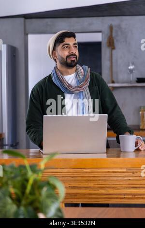 Mann, der hinter einer Holzinsel in der Küche steht, mit Mütze und Schal, mit Laptop neben dem Becher. Rustikal, modern, gemütlich, warm, Arbeitsbereich, Arbeitsplatte, Pflanze Stockfoto