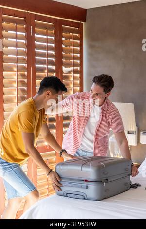 Zwei männliche Freunde stehen am Bett und packen graue Hartschalenkoffer in der Nähe von Holzläden. Mitreisende, Reise, Gepäck, Hotel, Schlafzimmer, lässig, sonnenbeleuchtet Stockfoto