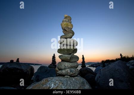 Ein ausgeglichener Steinkairn steht an einem felsigen Ufer in der Dämmerung, andere kleine steinkränze sind im Hintergrund zu sehen. Stockfoto