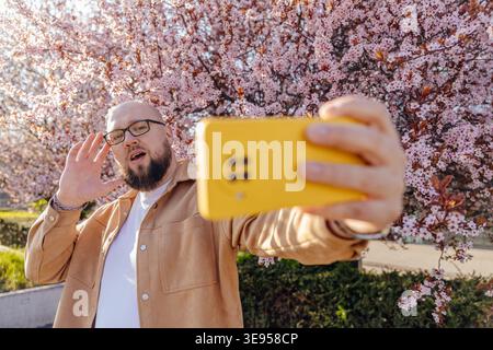 Der weiße Mann nimmt Selfie-Videos auf und winkt im Frühling in der Nähe des blühenden Kirschbaums Stockfoto
