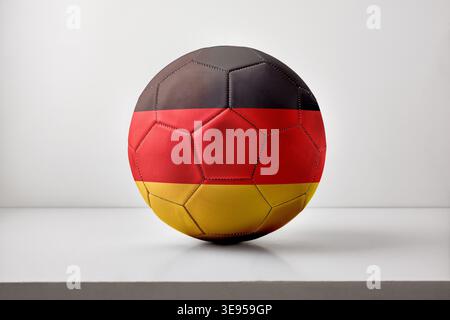 Ein Fußball, der mit den Flaggenfarben Deutschlands bemalt ist, sitzt auf einer sauberen weißen Oberfläche mit sanfter Beleuchtung und minimalem Hintergrund. Stockfoto