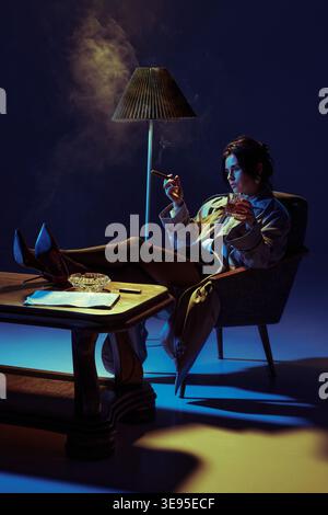 Elegante Frau, die Zigarre raucht und Whiskey im Sessel unter dramatischer Beleuchtung im Studio trinkt. Stockfoto