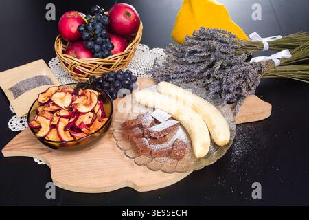 Auf einem Holzbrett werden frisches Obst wie Äpfel und Trauben, getrocknete Apfelscheiben, ein Schokoladendessert mit Puderzucker, Bananen und Bündel ausgestellt Stockfoto