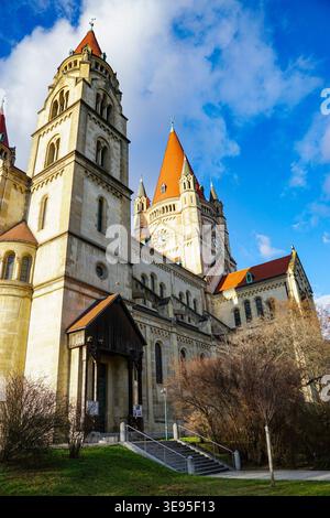 Historisches Kirchengebäude mit hohen Türmen und roten Ziegeldächern steht unter blauem Himmel mit Wolken neben Bäumen und Eingangstreppen. Stockfoto