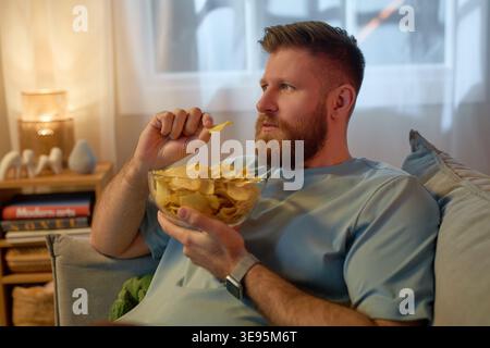 Erwachsener Mann isst Kartoffelchips, während er sich nach der Arbeit zu Hause entspannt Stockfoto