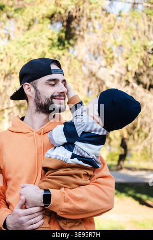 Vater hält den Jungen im Freien in den Armen, während das Kind sein Gesicht berührt und beide zeigen warme, liebevolle Ausdrücke. Erziehungsbegriff und Familiengabel Stockfoto
