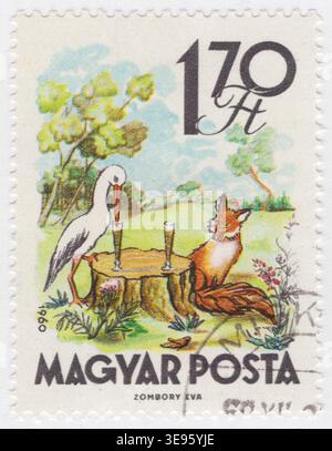 UNGARN - 1. Dezember 1960: 1,70 mehrfarbige Briefmarke zeigt Aesops Fabel "der Fuchs und der Storch". Die von der Künstlerin Eva Zombory geschaffene Szene zeigt die zweite Hälfte der Geschichte, in der sich der Storch gegen die Trickerei des Fuchses rächt. Ein Weißstorch und ein Rotfuchs sitzen an einem Tisch, der aus einem Baumstumpf auf einer grünen Wiese besteht. Zwei große, enghalsige Gläser werden auf dem Stumpf platziert. Der Storch trinkt mit seinem langen Schnabel leicht aus seinem Glas, während der Fuchs sitzt, ohne die Flüssigkeit zu erreichen. Der skurrile Aquarellstil fängt die moralische Lektion ein, andere so zu behandeln, wie Sie behandelt werden möchten Stockfoto