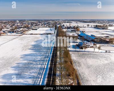 Hochwinkelansicht einer geraden Straße, die durch eine verschneite Winterlandschaft in Europa führt. Mit schneebedeckten Feldern, einer kleinen Stadt, Bauernhöfen Stockfoto