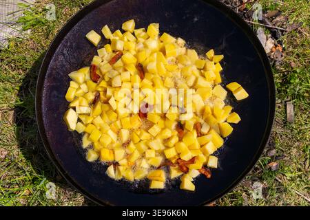 Gebratene Kartoffelwürfel in einer Pfanne im Freien kochen. Goldene Kartoffelwürfel, die in einer großen Pfanne im Freien mit brutzelndem Öl und Speckstückchen gebraten werden Stockfoto