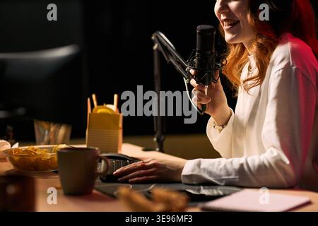 Junge Erwachsene kaukasische Frau, die in ein Podcast-Mikrofon spricht, während sie Inhalte am Schreibtisch streamt und Live-Medien für die Publikumsinteraktion erstellt. Ideal für kreative Wirtschaft, Blogging und Online-Übertragung Stockfoto