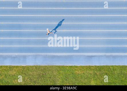 Luftaufnahme eines einsamen Läufers, der auf einer blauen Sportstrecke joggt, mit weißen Spurlinien und einem langen Schatten auf der Oberfläche neben einem grünen Streifen Stockfoto