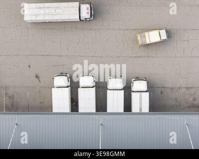 Aus der Vogelperspektive eines Logistikzentrums mit weißen Lkws, die an Laderampen geparkt sind, und einem Fahrzeug, das sich über gerissenen Asphalt in Taiwan bewegt. Stockfoto