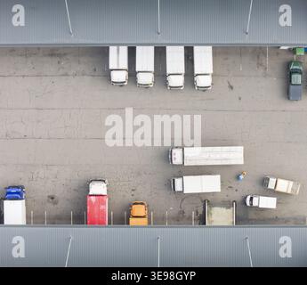 Aus der Vogelperspektive eines belebten Logistikzentrums mit farbenfrohen Lkw und Lieferwagen, die zwischen grauen Wellpappendächern auf einer gerissenen Asphaltsurfe geparkt sind Stockfoto