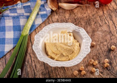 Cremiger Hummus-Dip in dekorativer weißer Schüssel. Frische Kichererbsen, Knoblauch, grüne Zwiebeln, Tomaten kunstvoll auf rustikalem Holzhintergrund angeordnet Stockfoto