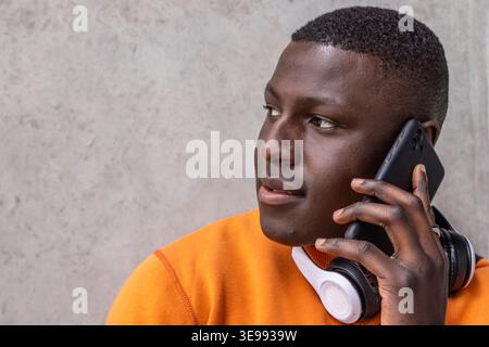 Afroamerikaner mit schwarzem Telefon am Ohr, orangefarbenem Sweatshirt, Headset gegen graue Wand Stockfoto