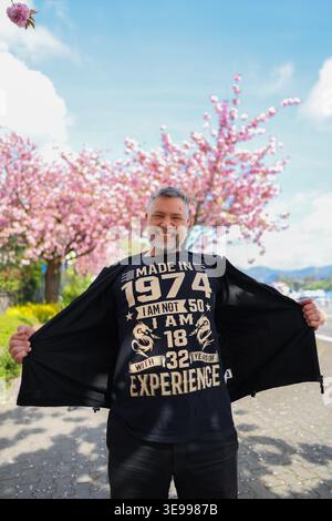 Lächelnder Mann im Frühlingspark unter blühenden Kirschbäumen mit lustigem Geburtstags-T-Shirt, Lifestyle-Porträt mit positiver Stimmung. Stockfoto