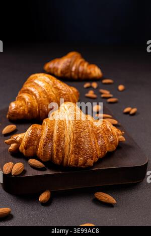 Frisch gebackene goldene Croissants mit knuspriger Konsistenz, serviert neben verstreuten Mandeln auf einem dunklen Holzschneidebrett, serviert mit einem köstlichen Frühstück Stockfoto