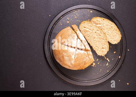 Rustikales Brot mit frisch gebackenem Brot mit Kreuzschlitz und zwei Scheiben, auf einem dunklen runden Schneidebrett mit Krümeln, gegen einen dunklen Kontrast Stockfoto