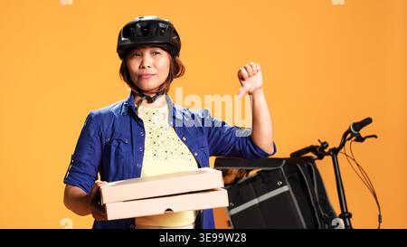 Nahaufnahme eines Teilzeit-Kuriers, der Pizzakartons zum Mitnehmen hält und das Daumen-nach-unten-Schild zeigt. Fahrradfahrer, der eine missbilligende Fingerschüttelbewegung macht, nein sagt, isoliert über Studiohintergrund Stockfoto