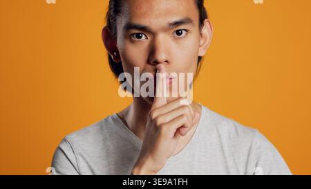 Privater asiatischer Typ, der stille Schilder vor der Kamera macht, Stummschaltungssymbole macht, um geheime und vertrauliche Nachrichten zu bewahren. Junge Person, die im Studio Privatsphäre und Geheimhaltung wahrt, mit Informationen vorsichtig ist. Stockfoto