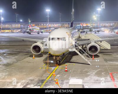 Riga, Lettland - 01. Februar 2026: : Flugzeug, das nachts auf dem Asphalt geparkt ist, mit einer Treppe, einem Flughafenterminal und zusätzlichen Flugzeugen in t Stockfoto