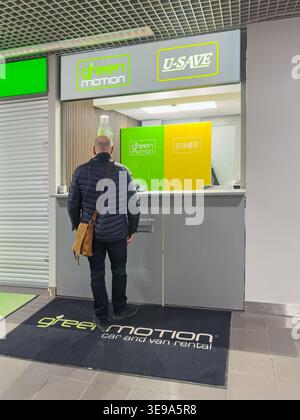 Riga, Lettland - 03. April 2026: Mann in dunkler Jacke nähert sich Green Motion Autovermietung mit U-SAVE-Schild in einer modernen Innenumgebung Stockfoto