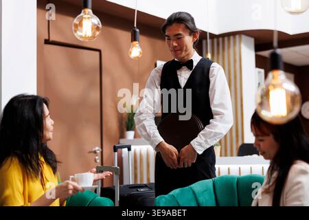 Asiatische Frau mittleren Alters, die während des Urlaubs Kaffee von einem lächelnden männlichen Concierge im Foyer des gemütlichen Resorts erhält. Mitarbeiter, die neue Kunden in der komfortablen Lounge bestellen Stockfoto