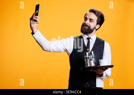 Selbstbewusster Kellner, der Selfies im Studio macht, mit der Smartphone-Kamera Bilder von ihm mit dem Tablett in der Hand aufnimmt. Junger Mann, der als Butler arbeitet und Fotos vor gelbem Hintergrund macht. Stockfoto
