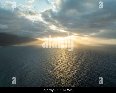 Aus der Vogelperspektive auf den Atlantischen Ozean mit goldenen Sonnenstrahlen, die durch dramatische Wolken über der zerklüfteten Küste Madeiras, Portugal, brechen. Stockfoto