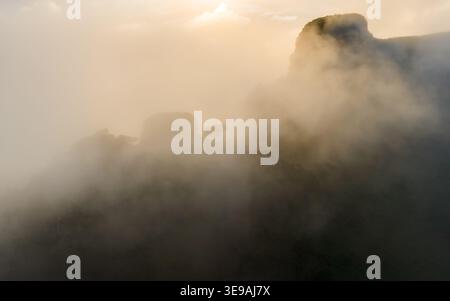 Blick aus der Vogelperspektive auf die alten Lorbeerbäume des Fanal-Waldes, umgeben von dickem Morgennebel mit goldenem Sonnenlicht, das durch die Wolken in Seixal, verrückt, bricht Stockfoto