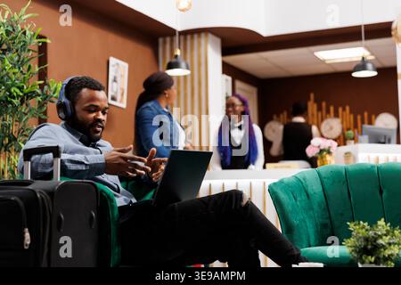 Fokussierter afroamerikanischer Geschäftsmann mit Kopfhörern, der Videoanrufe mit Kunden auf einem Laptop hält, während er mit Koffer in der Hotellobby sitzt. Unternehmer, der auf Reisen an Videogesprächen teilnimmt Stockfoto