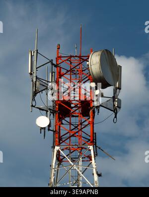 Ein Telekommunikationsturm mit mehreren Antennen und einer großen Schüssel, in Rot und weiß gestrichen, steht vor einem blauen Himmel mit verstreuten Wolken. Stockfoto