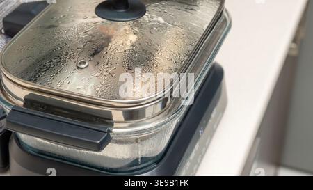 Glasbehälter mit Deckel mit Kondenswasser auf schwarzem Induktionskochfeld in der Küche. Hausmannskochprozess, Dämpfen von Speisen, Abdeckung Stockfoto