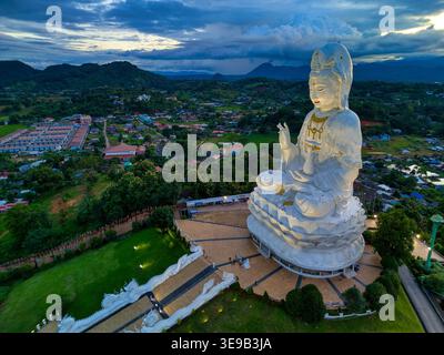 Blick aus der Vogelperspektive auf die riesige weiße Guan Yin Statue im Wat Huay Pla Kang Tempelkomplex, umgeben von üppigen grünen Hügeln und einem lokalen Dorf unter einem dramatischen Stockfoto