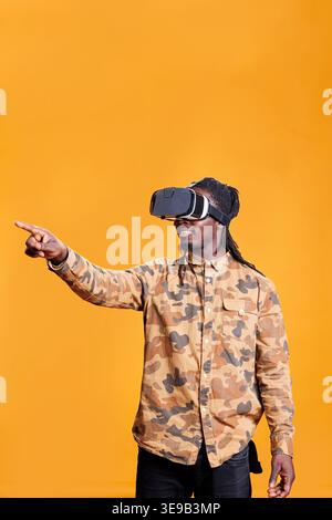 Selbstbewusste Person, die eine vr-Brille für 3D-Simulation trägt und virtuelle Realität mit interaktivem Headset genießen kann. Fröhlicher afroamerikanischer Mann, der Spaß mit Brillen im Studio auf gelbem Hintergrund hat Stockfoto