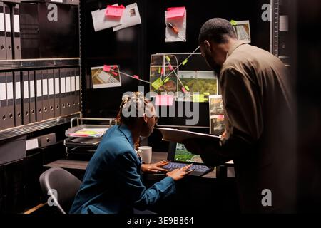 Detectives analysieren Falldateien und suchen Informationen auf dem Laptop, während sie spät in der Agentur arbeiten. Die Ermittler lesen csi-Berichte und überprüfen Tatortfotos auf dem Computerbildschirm Stockfoto
