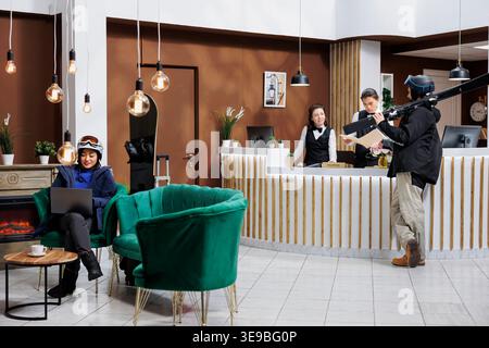 Das Bild zeigt eine asiatische Frau mit einem Snowboard, das mit einem Laptop auf dem Sofa sitzt, während ein junger Mann mit Skiern an der Rezeption steht. Besucher in Schneekleidung und Wintersportausrüstung in der Hotellobby zum Check-in. Stockfoto