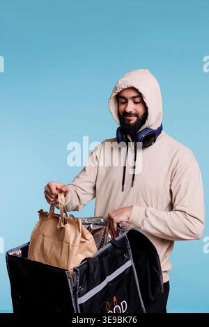Arabischer Essenslieferant in der Haube, der ein Paket mit Mittagessen aus dem Rucksack nimmt. Junger bärtiger Mann mit beigefarbenem Kapuzenpullover, der Papiertüte mit Take-away-Schale und Thermotasche hält Stockfoto