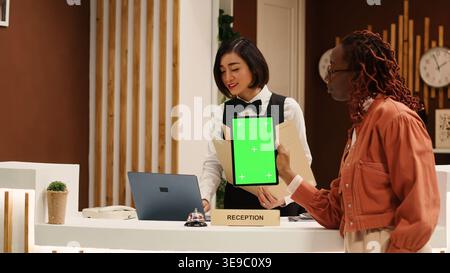Müde Touristen halten Chroma Key Green Screen Mock Up Tablet an der Hotelrezeption nach einem langen Flug und warten darauf, dass die Zimmerzahlung bearbeitet wird. Gast im Hotelfoyer, der eincheckt Stockfoto