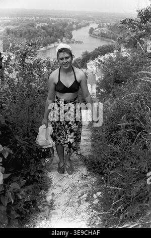 Historischer Archivblick von Slavyanogorsk (heute Sviatohirsk) aus dem Jahr 1956 mit Valentina Timofeevna Kravelis, der einen Sommerurlaub genießt. Die Szene zeigt die junge Frau, die einen steilen, kalkhaltigen Waldweg mit dunklem Bikinioberteil und einem Rock mit Blumenmuster hinaufgeht, mit dem Tal des Flusses darunter. Dieses nostalgische Porträt aus der Mitte des zwanzigsten Jahrhunderts zeigt Outdoor-Freizeit, persönliche Mode und die natürliche Schönheit der Sowjetzeit, bewahrt das historische tägliche Leben und die einzelnen Menschen der Region und spiegelt den friedlichen Donbass wider. Stockfoto