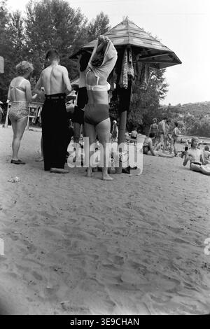 Historischer Archivblick von Slavyanogorsk (heute Sviatohirsk) aus dem Jahr 1956 mit Sommerurlaub entlang der Sandufer des Siwerskyi Donets. Die Szene zeigt Urlauber, die sich unter einem hölzernen Sonnenschirm entspannen und ihre Kleidung wechseln und auf einem schattigen Sandweg spazieren gehen, der von hohen, grünen Bäumen umgeben ist. Dieser nostalgische Moment in der Mitte des zwanzigsten Jahrhunderts beleuchtet die Freizeitaktivitäten im Freien, die ungezwungene Strandkultur und die natürliche Umgebung der Sowjetzeit und bewahrt das historische tägliche Leben der Region und spiegelt den friedlichen Donbass wider. Stockfoto