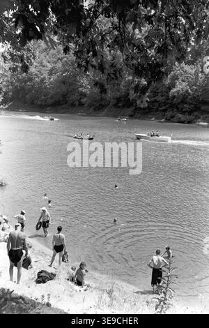 Historischer Blick aus dem Jahr 1956 auf Slavjanogorsk (heute Sviatohirsk) mit Outdoor-Freizeitaktivitäten auf dem malerischen Fluss Siwerskyi Donets. Die Szene zeigt Einheimische, die im ruhigen Wasser schwimmen und sich am Sandstrand entspannen, während mehrere hölzerne Ruderboote und Motorboote an den bewaldeten Ufern vorbeigleiten. Diese nostalgische Landschaft aus der Mitte des zwanzigsten Jahrhunderts unterstreicht die natürliche Schönheit und die lebendige Sommerurlaubskultur der Sowjetzeit, die die historische Umgebung und das tägliche Leben der Region bewahrt und den friedlichen Donbass widerspiegelt. Stockfoto