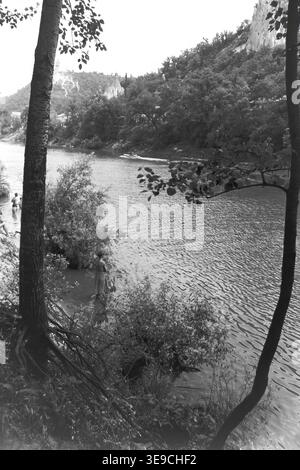 Historischer Archivblick von Slavyanogorsk (heute Sviatohirsk) aus dem Jahr 1956 mit Blick auf den malerischen Fluss Siwerskyi Donets, der von dunklen Baumsilhouetten eingerahmt wird. Die Szene fängt kleine Ruderboote und eine Passagier-Flussbahn auf dem Wasser ein, während die majestätische Kreidekirche St. Nikolaus auf der Klippe im Hintergrund aufsteigt. Diese nostalgische Landschaft aus der Mitte des zwanzigsten Jahrhunderts hebt die natürliche Schönheit und das architektonische Erbe der Sowjetzeit hervor und bewahrt die historische Umgebung der Region und spiegelt den friedlichen Donbass wider. Stockfoto