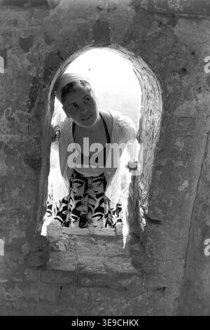 Historischer Archivblick von Slavyanogorsk (heute Sviatohirsk) aus dem Jahr 1956 mit Valentina Timofeevna Kravelis bei der Erkundung der historischen Ruinen. Die Szene fängt die junge Frau ein, die sich durch ein rustikales, bogenförmiges Steinfenster mit Klippenstruktur lehnt, gekleidet mit Bikinioberteil, gestreiftem Hemd und gemustertem Rock, während sie nach außen blickt. Dieses nostalgische Porträt aus der Mitte des zwanzigsten Jahrhunderts zeigt persönliche Reisen, Outdoor-Tourismus und architektonisches Erbe der sowjetischen Ära, wobei die historischen Menschen der Region erhalten bleiben und den friedlichen Donbass widerspiegeln. Stockfoto