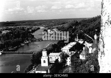 Historischer Archivblick von Slavyanogorsk (heute Sviatohirsk) aus dem Jahr 1956 mit einem atemberaubenden Panorama auf den Fluss Siwerskyi Donets. Die Szene fängt eine hohe Perspektive mit Blick auf die historischen Gebäude von Svyatogorsk Lavra mit ihren charakteristischen Kuppeln, einer Fußgängerbrücke über das Wasser und das weite bewaldete Tal ein. Diese nostalgische Landschaft aus der Mitte des zwanzigsten Jahrhunderts hebt die architektonische Pracht, die natürliche Geographie und das religiöse Erbe der Sowjetzeit hervor und bewahrt die historische Umgebung, die den friedlichen Donbass widerspiegelt. Stockfoto