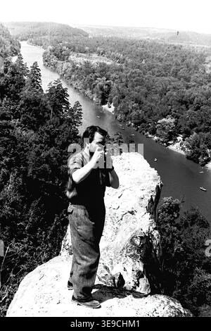 Historischer Archivblick von Slavyanogorsk (heute Sviatohirsk) aus dem Jahr 1956, der einen Besucher zeigt, der die atemberaubende Landschaft dokumentiert. Die Szene zeigt einen Mann, der auf einem zerklüfteten Felsvorsprung hoch über dem Fluss Siverskyi Donets steht und eine Vintage-Kamera hält, um den gewundenen Wasserweg und die ausgedehnten dichten Sommerwälder zu fotografieren. Dieser nostalgische Moment in der Mitte des zwanzigsten Jahrhunderts beleuchtet den Tourismus, die Amateurfotografie und die dramatische natürliche Schönheit der Sowjetzeit, die die historische Umgebung der Region bewahrt und den friedlichen Donbass widerspiegelt. Stockfoto