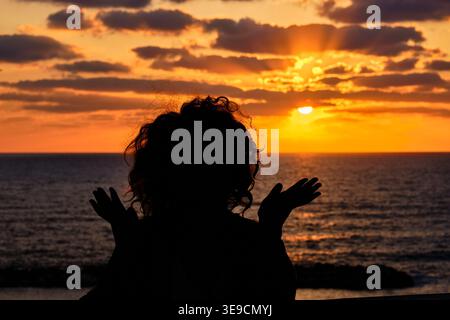 Silhouette einer Frau mit lockigem Haar, die einen goldenen Sonnenuntergang über dem Meer beobachtet. Friedlicher und emotionaler Moment mit Ozeanhorizont und warmem Abendlicht. Stockfoto