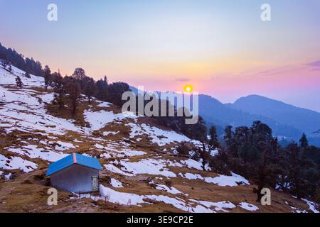 Eine kleine Hütte mit blauem Dach befindet sich auf einem schneebedeckten Berghang bei Sonnenuntergang in der Nähe des Tungnath Tempels in Uttarakhand, Indien. Die Landschaft ist gespickt mit Bäumen und Stockfoto